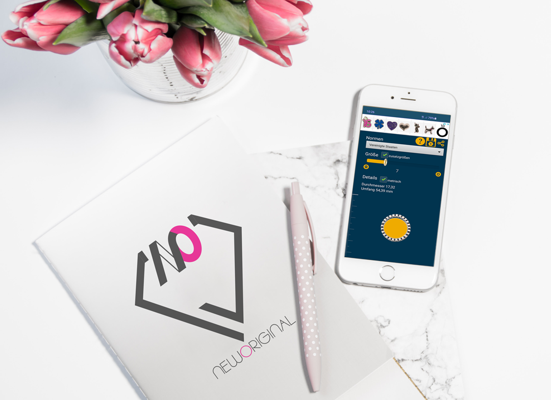 Smartphone mit App zur Ringgrößenbestimmung liegt auf einem Schreibtisch neben einem Notizheft, einem Stift und einer Vase mit Tulpen. 